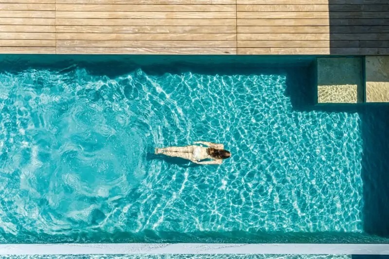 Bovenaanzicht van zwemmende vrouw in uxe living pool met houten vlonders en trapje naar boven.