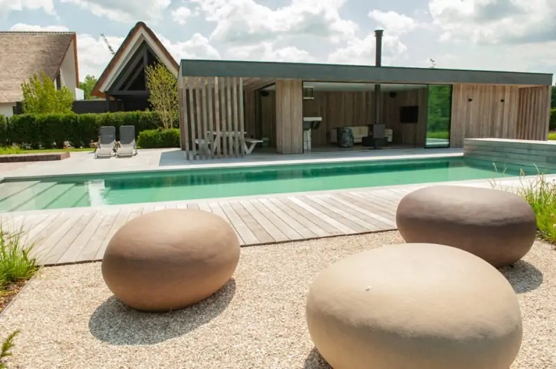 Strakke living pool bij moderne villatuin met houten poolhouse, loungeterras en helder natuurlijk zwemwater