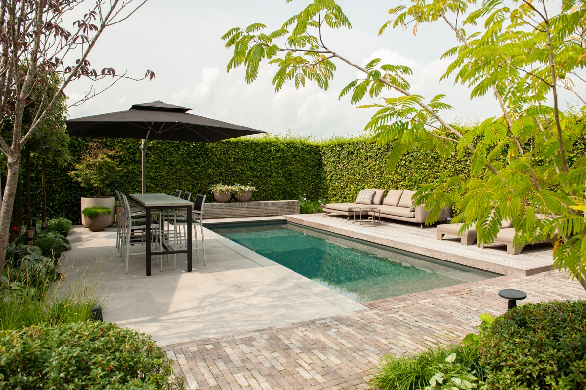 Strakke living pool ingebed in een tuin met natuurstenen terras, bakstenen bestrating, eethoek en loungehoek omgeven door groene hagen.