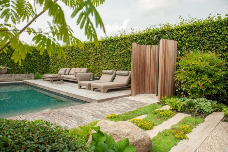 Living pool met houten buitendouche, ligbedden en loungehoek, omringd door groene hagen en uiteenlopende beplanting.