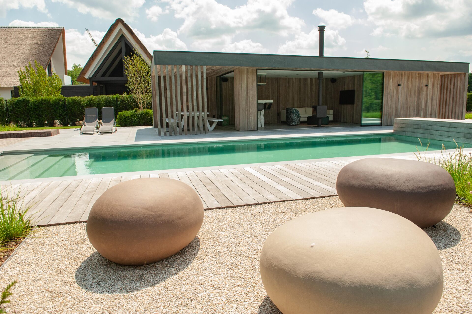 Strakke living pool met houten vlonder en modern buitenverblijf met lounge, eethoek en haard.