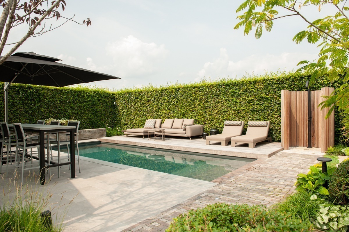 Strakke living pool met terras, eethoek, loungeplek en houten buitendouche in een besloten groene tuin.
