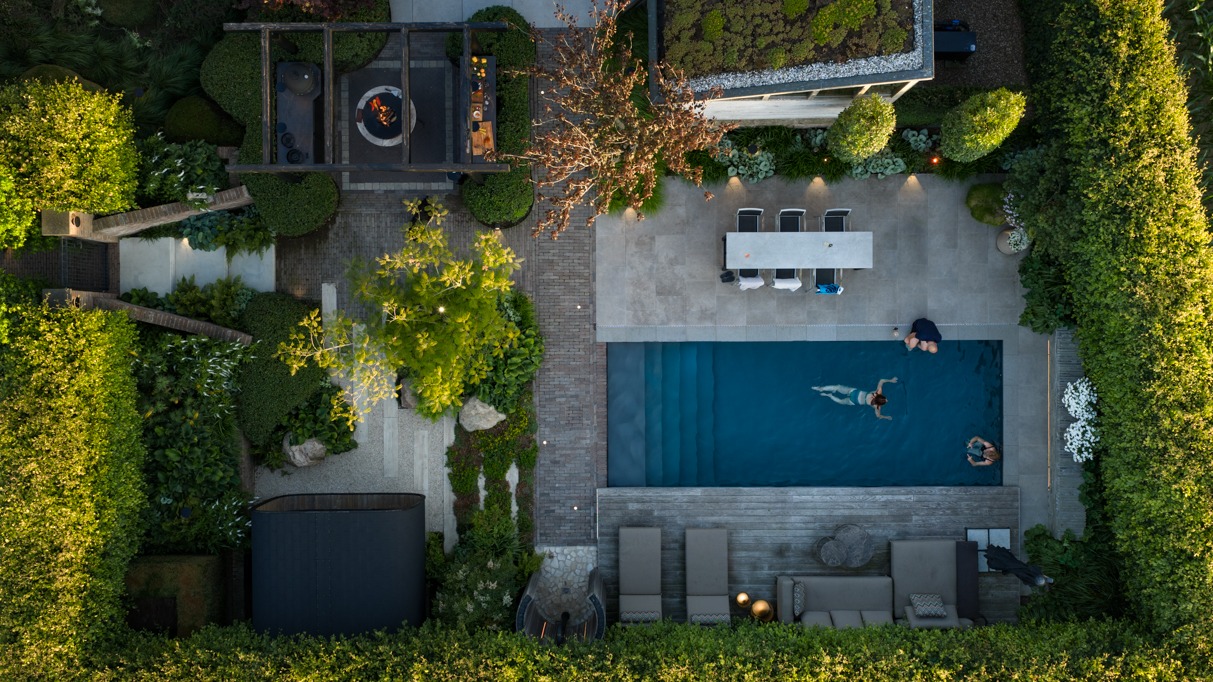 Bovenaanzicht van een moderne achtertuin met een rechthoekig blauw zwembad waarin iemand zwemt. Rondom het zwembad liggen een houten terras met ligbedden en een hoekbank, een betegeld terras met een lange eettafel, en een houten pergola met vuurschaal. Het geheel is omheind met groene heggen.