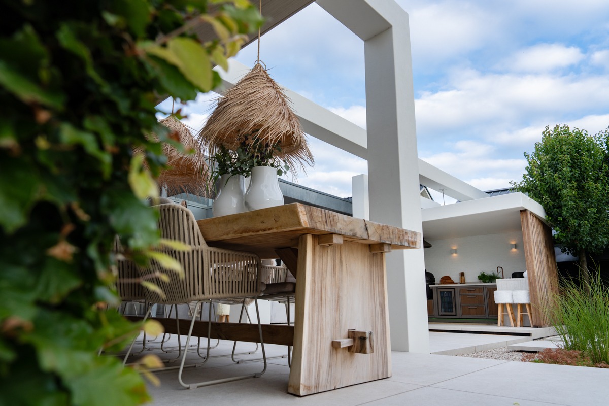Een robuuste houten eettafel met gevlochten stoelen en bohemian rieten lampen op een strak betegeld terras onder een witte pergola. Op de achtergrond is een overdekte buitenkeuken te zien met houten afwerking, een bar met krukken en inbouwapparatuur.