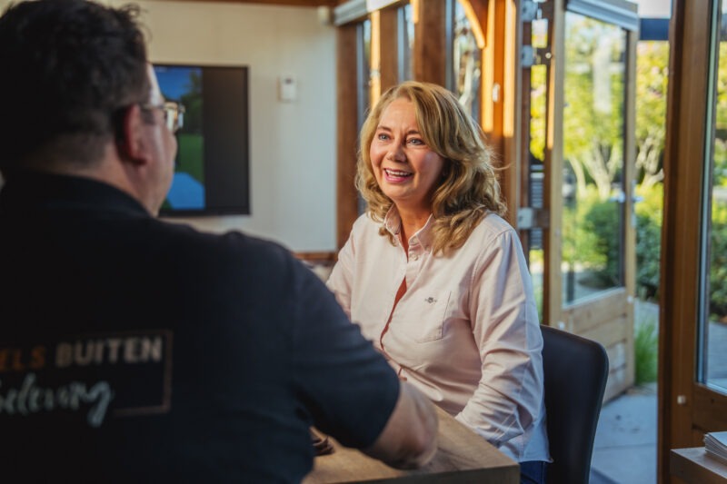 Een blonde vrouw in een lichtroze overhemd zit glimlachend aan een houten tafel in gesprek met een adviseur op de voorgrond. Het gesprek vindt plaats in een lichte ruimte met grote glazen deuren die uitzicht bieden op een groen buitenbereik.