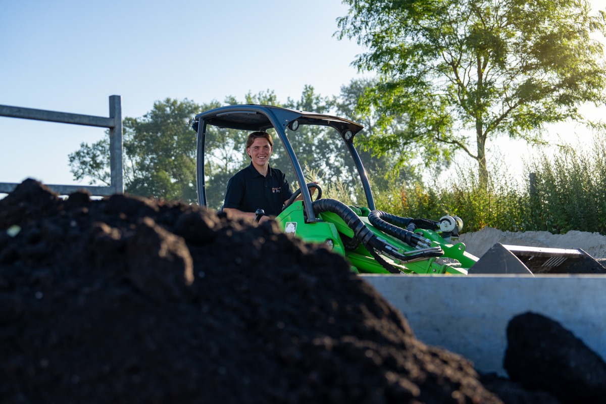Een jonge man zit glimlachend op een groene kniklader tijdens grondwerkzaamheden. Op de voorgrond ligt een grote hoop zwarte tuinaarde, met op de achtergrond bomen en een heldere blauwe lucht.