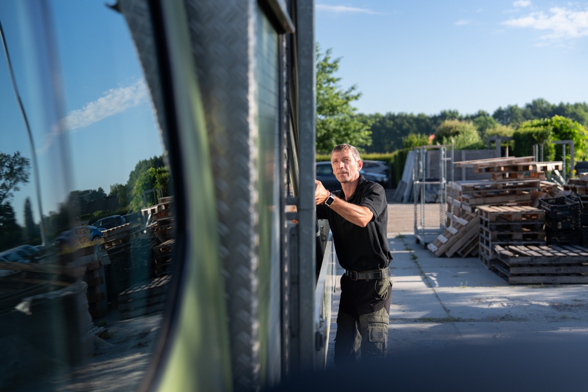 Een man in zwarte werkkleding kijkt geconcentreerd terwijl hij werkzaamheden uitvoert bij de zijkant van een werkbus. Op het buitenterrein staan opgestapelde houten pallets en op de achtergrond is een groene omgeving onder een heldere lucht te zien.