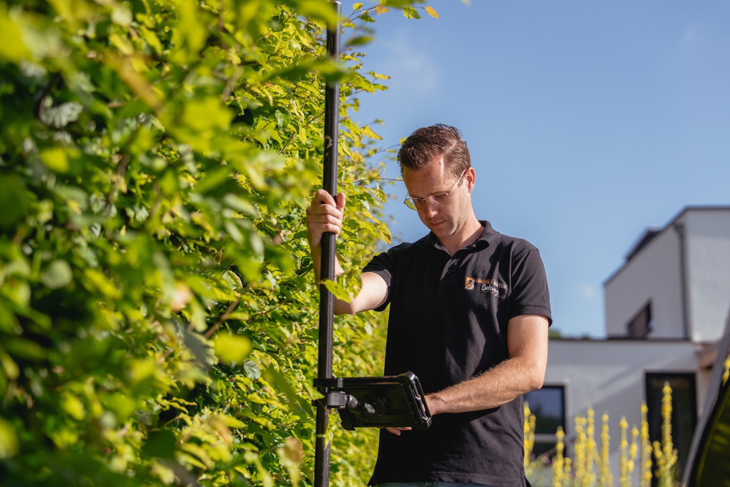 Een man met een bril en een zwarte polo gebruikt een GPS-inmeetstok met een digitaal scherm bij een volle groene heg. Op de achtergrond is een moderne witte woning te zien onder een heldere blauwe lucht.