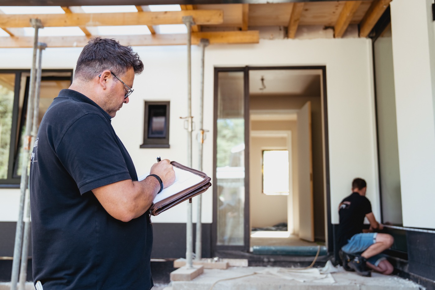 Een man met een bril en zwarte polo maakt aantekeningen in een schrijfmap op de voorgrond. Op de achtergrond is een modern huis in aanbouw te zien met stempels onder een overkapping, waar een tweede vakman gehurkt werkzaamheden bij de gevel uitvoert.