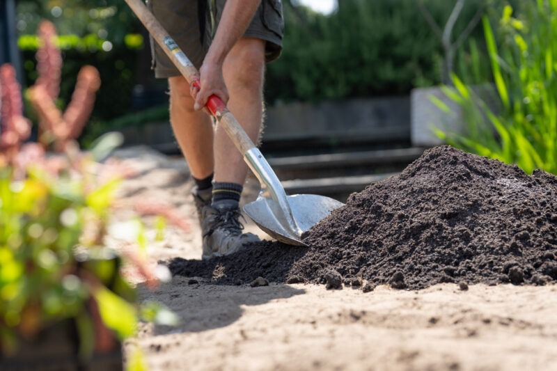 Hovenier schept met een spade zwarte grond op. Op de voorgrond staan wat planten klaar om aangeplant te worden.