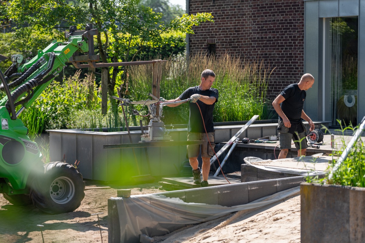 Twee hoveniers leggen een terras aan achter een woning. Eén man bedient een vacuümtilapparaat aan een groene kniklader om een grote zware tegel te verplaatsen, terwijl zijn collega op de achtergrond bij de gevel bezig is met werkzaamheden.