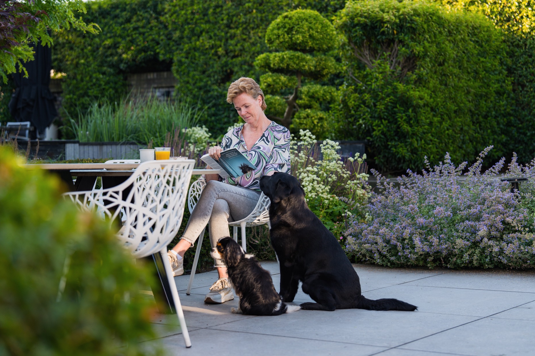 Een vrouw zit op een witte designstoel aan een tuintafel een boek te lezen. Naast haar op het terras zitten een grote zwarte hond en een kleine spaniel. De achtergrond bestaat uit een rijk beplante tuin met paarse en witte bloemen, vormsnoei en dichte groene heggen.