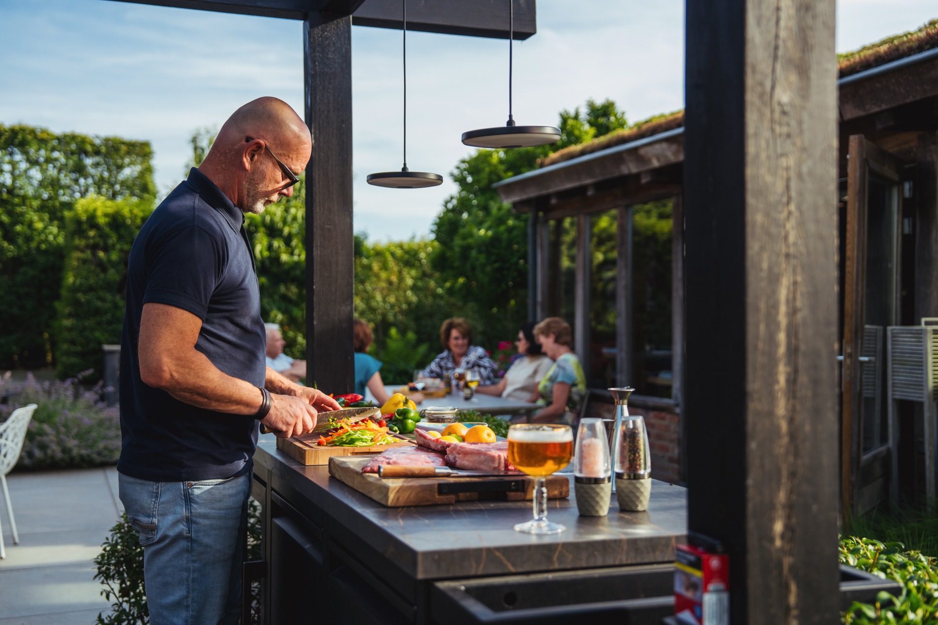 Een man met een bril en donker shirt snijdt groenten op een houten snijplank aan het kookeiland van een moderne buitenkeuken. Op het aanrecht liggen vlees, groenten en staat een glas bier. Op de achtergrond zit een groep mensen aan een eettafel in de tuin bij een bijgebouw met een groendak.