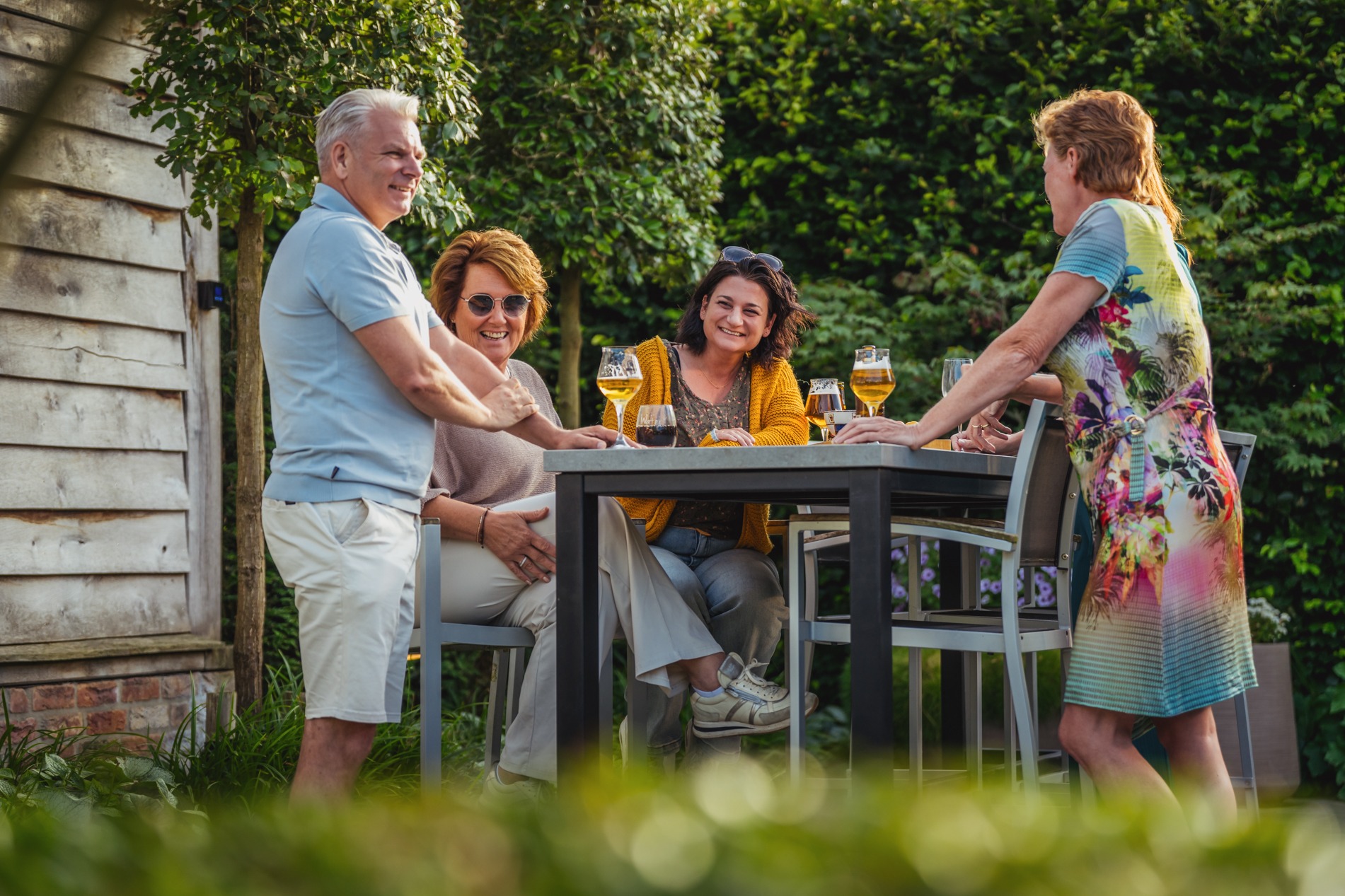 Een gezelschap van vier volwassenen staat en zit rond een hoge eettafel met glazen bier en wijn. Ze praten en lachen met elkaar in een zonnige tuin met een dichte groene heg en een houten bijgebouw op de achtergrond.