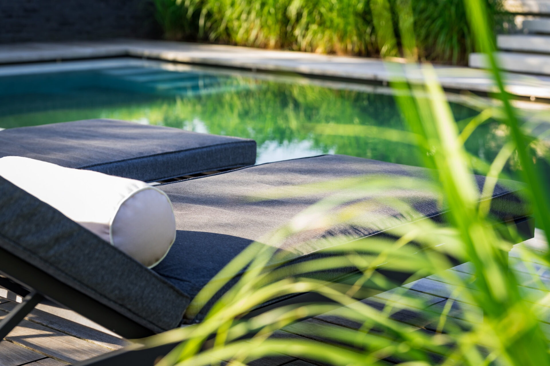 Luxe ligbedden op een houten vlonder naast helder water van een living pool, omringd door siergrassen.
